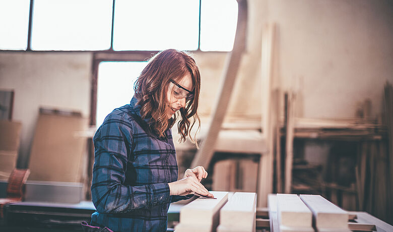 In einer Werkstatt schleift eine junge Frau große Kanthölzer ab