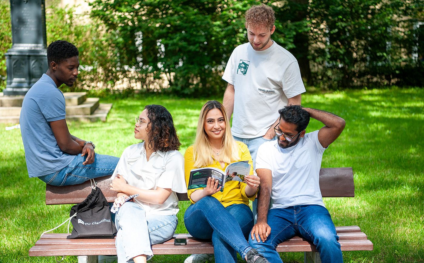 Fünf Studierende sitzen auf einer Bank im Grünen auf dem Campus
