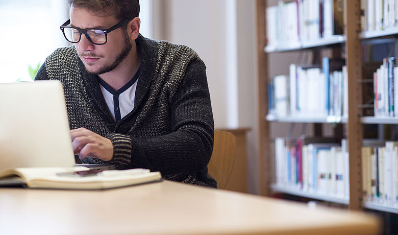 In einer Bibliothek arbeitet ein junger Mann konzentriert an seinem Laptop 
