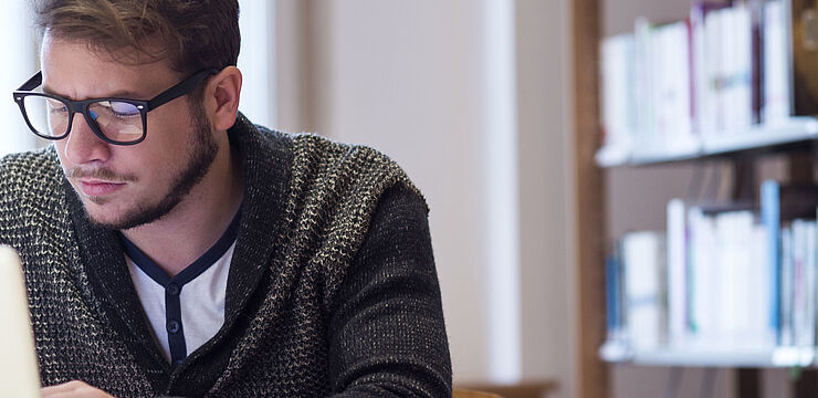 In einer Bibliothek arbeitet ein junger Mann konzentriert an seinem Laptop 