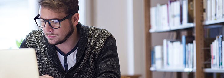 In einer Bibliothek arbeitet ein junger Mann konzentriert an seinem Laptop 