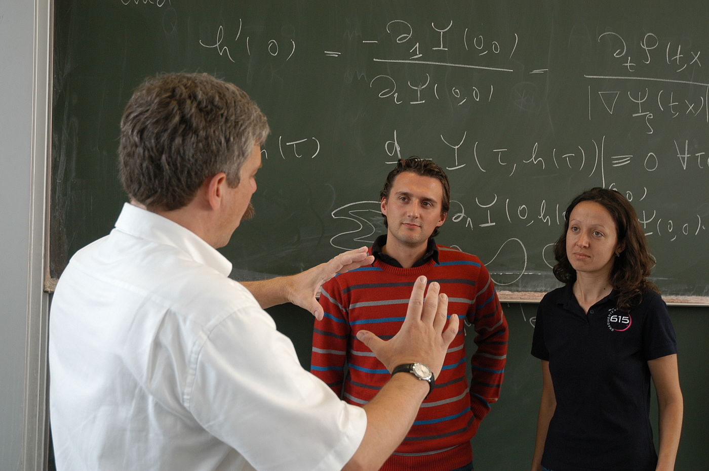 Ein Lehrender erklärt zwei Studierenden etwas vor einer Tafel voller mathematischer Gleichungen