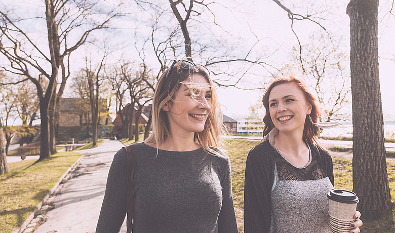 Zwei junge Frauen spazieren fröhlich mit Coffee to go durch einen Park und unterhalten sich