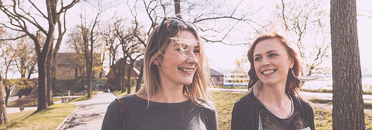 Zwei junge Frauen spazieren fröhlich mit Coffee to go durch einen Park und unterhalten sich