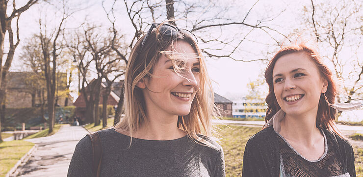 Zwei junge Frauen spazieren fröhlich mit Coffee to go durch einen Park und unterhalten sich
