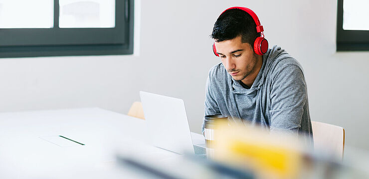 In einer Bibliothek arbeitet ein junger Mann mit Kopfhörern konzentriert an seinem Laptop 