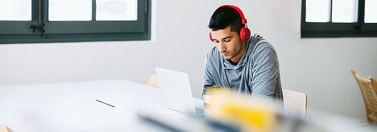 In einer Bibliothek arbeitet ein junger Mann mit Kopfhörern konzentriert an seinem Laptop 