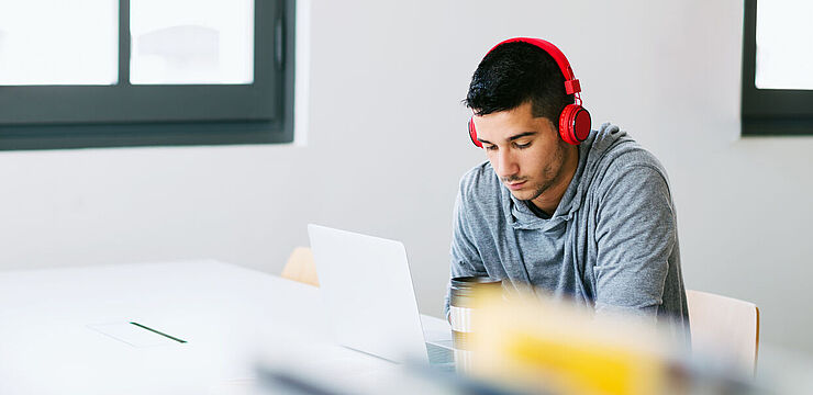 In einer Bibliothek arbeitet ein junger Mann mit Kopfhörern konzentriert an seinem Laptop 