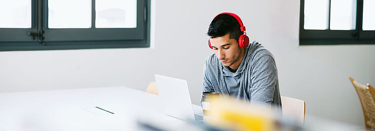 In einer Bibliothek arbeitet ein junger Mann mit Kopfhörern konzentriert an seinem Laptop 