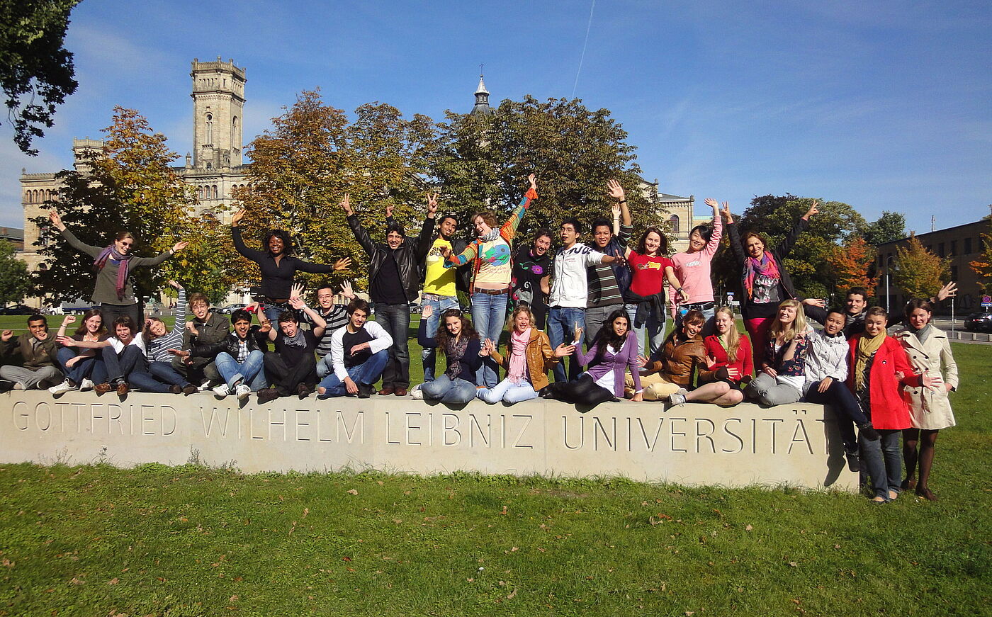 Viele Studierende winken vom Schild der Universität Hannover vor dem Hauptgebäude