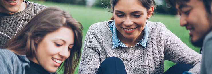 Auf einer Wiese sitzen mehrere junge Menschen zusammen und betrachten etwas auf einem Tablet-PC